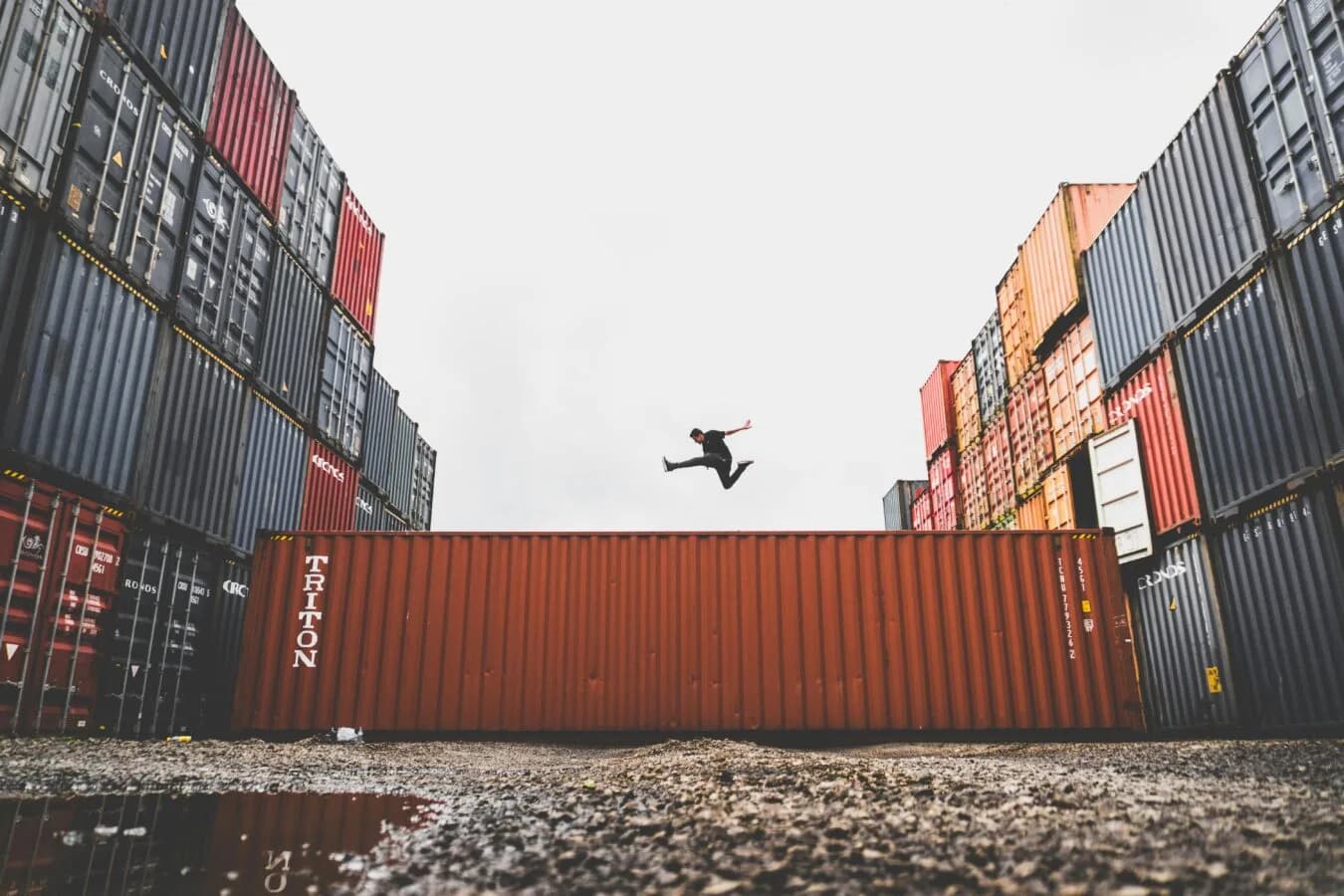 Logistikhof mit gestapelten Frachtcontainern und einer Person, die über einen roten Container springt, Lufthansa Cargo Stichwort sichtbar