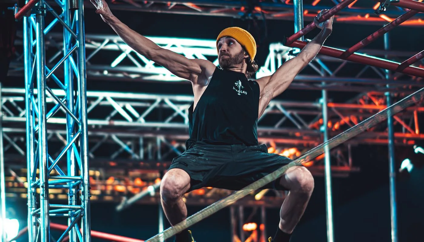 Athlet in gelber Mütze und schwarzem Tanktop klettert an Metallgerüst und Hindernisstreben bei einem Parcours- oder Ninja-Wettkampf