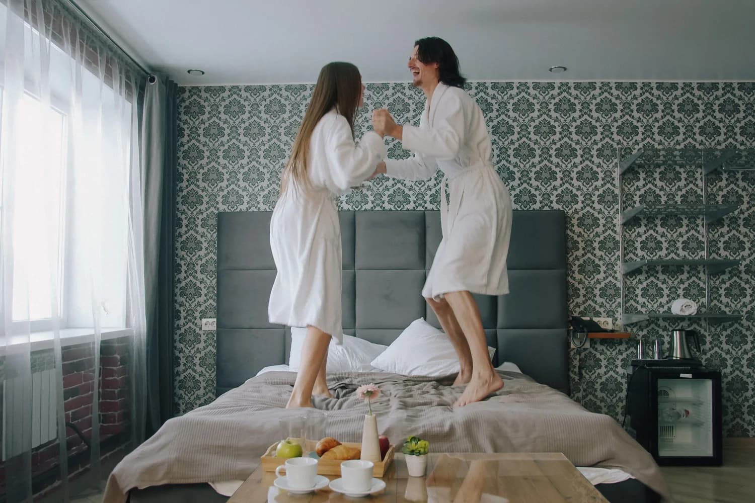 Zwei Personen in Bademänteln springen lachend auf einem Hotelbett, davor ein Tisch mit Frühstück