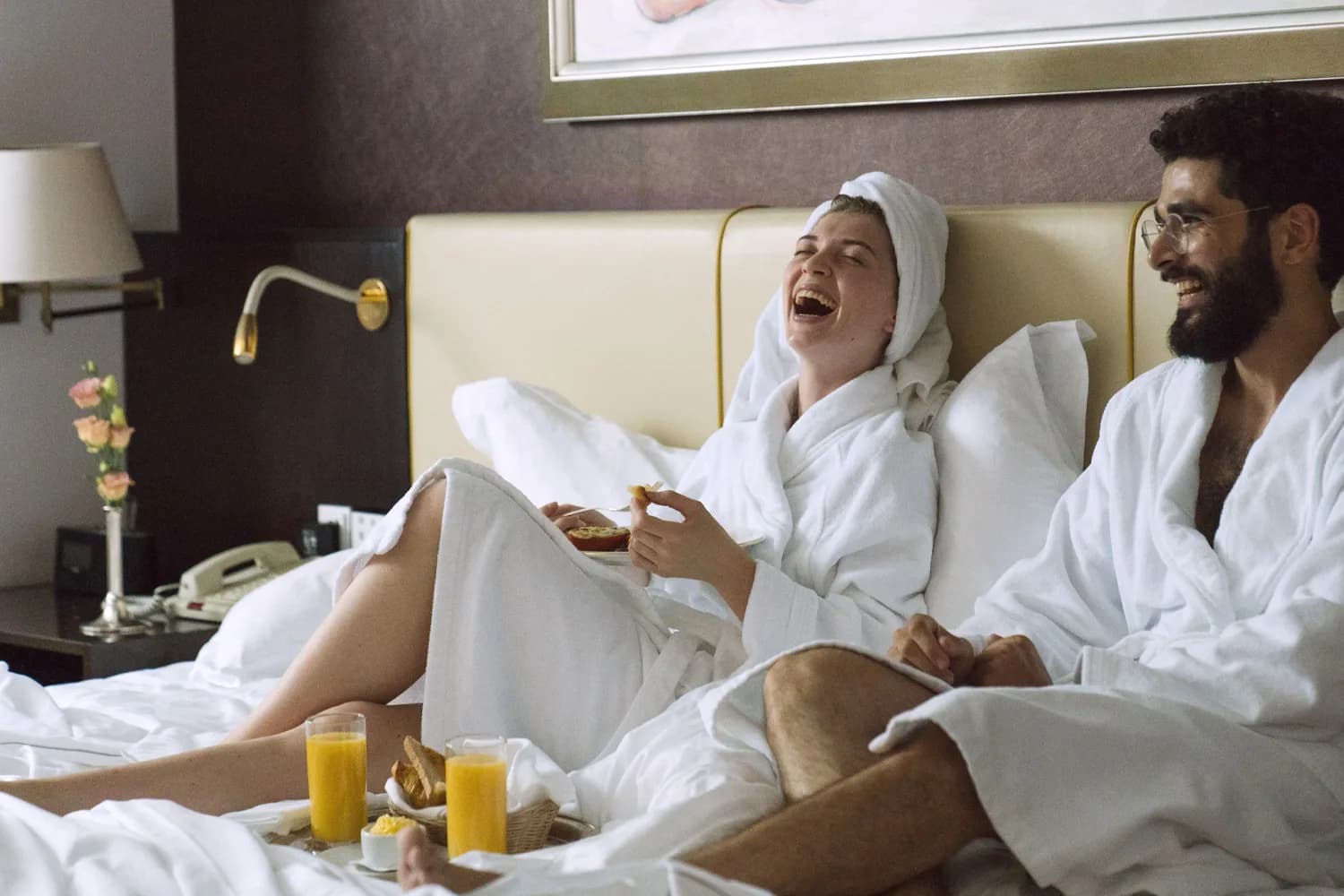Paar in Bademänteln lacht im Hotelbett beim Frühstück mit Orangensaft und Gebäck