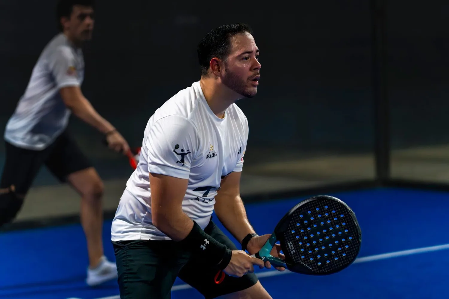 Zwei Padel-Spieler in weißen Trikots auf blauem Court, Vordergrundspieler in Bereitschaft mit Schläger für Rückhand, Team-Matchszene