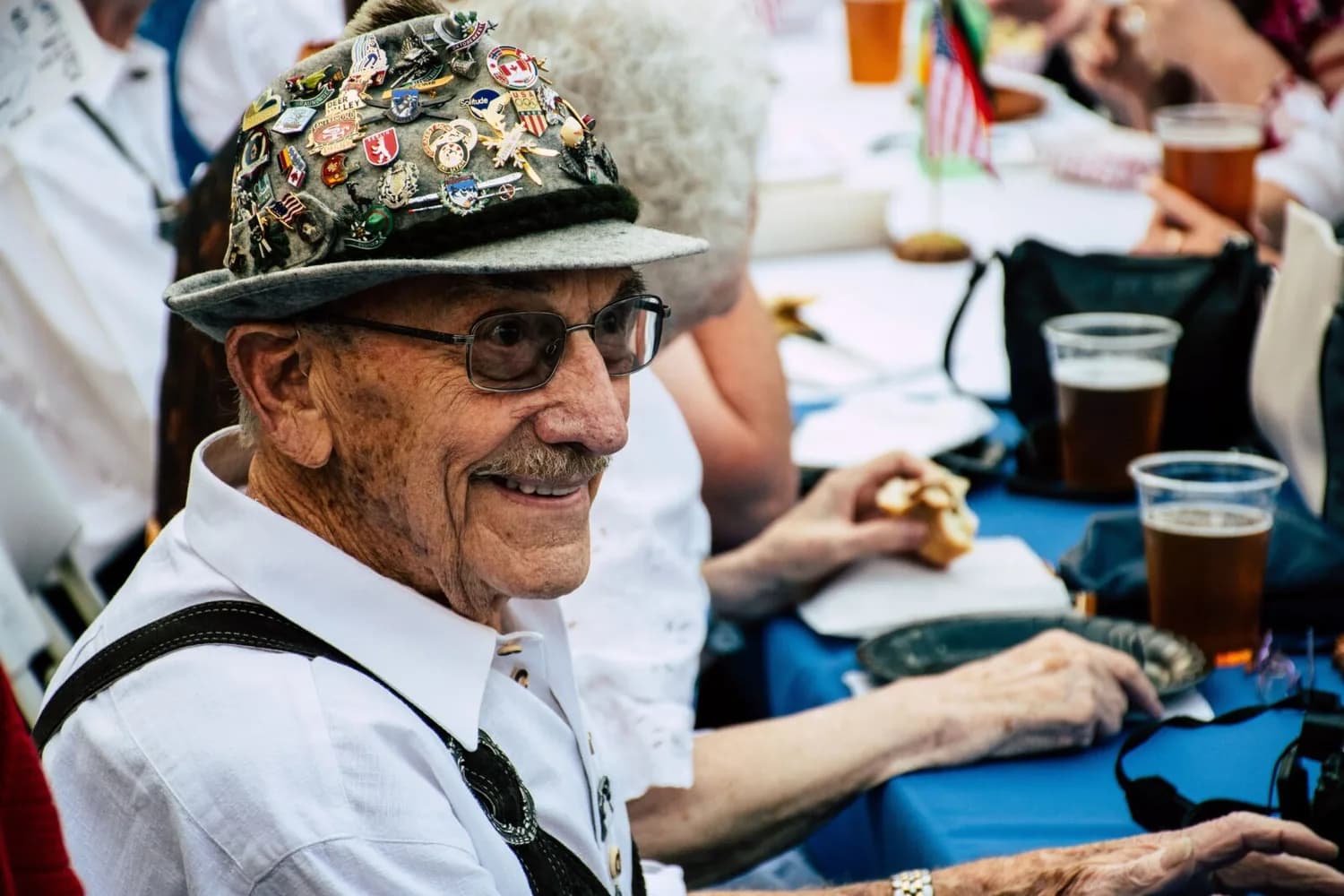 Älterer Mann in Trachtenhut mit Sammelanstecknadeln und Lederhosenträgern sitzt bei Volksfesttisch mit Bier und Brotzeit