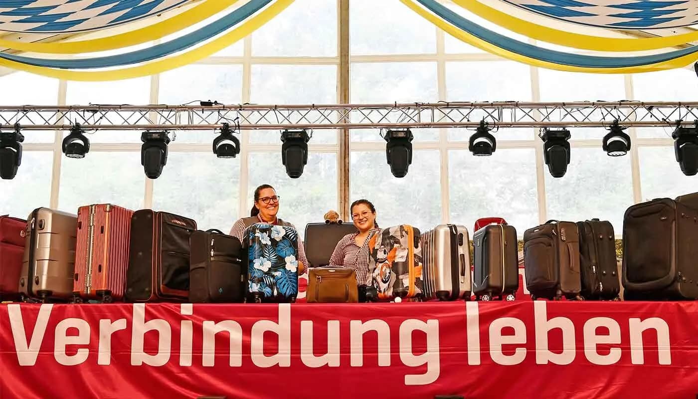 Zwei lächelnde Mitarbeiterinnen hinter aufgereihten Fundkoffern und Reisetaschen bei Fundsachenversteigerung am Flughafen München, Koffer verschiedener Farben und Größen auf Bühne mit Beleuchtung und rotem Banner "Verbindung leben"