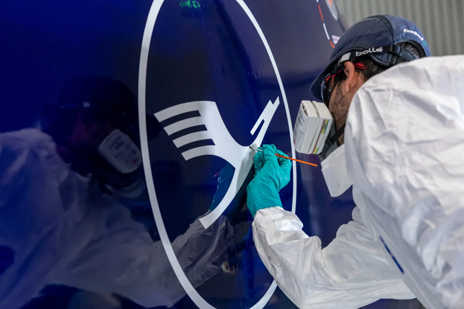 Techniker in Schutzanzug und Atemschutzmaske retuschiert Lufthansa-Logo auf dunkelblauer Flugzeugheckseite mit feinem Pinsel