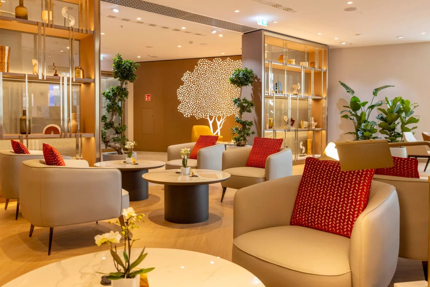 Luxuriöse Emirates-Lounge am Flughafen München mit bequemen Sesseln, roten Kissen, Kaffee- und Beistelltischen, Pflanzen und dekorativem Wandbaum – entspannter Wartebereich für Reisende