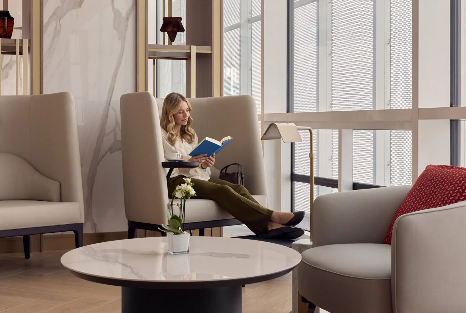 Frau liest Buch in ruhiger Lounge-Ecke mit hohen Polstersesseln, Beistelltisch, Lampe und Ausblick auf Fensterfront