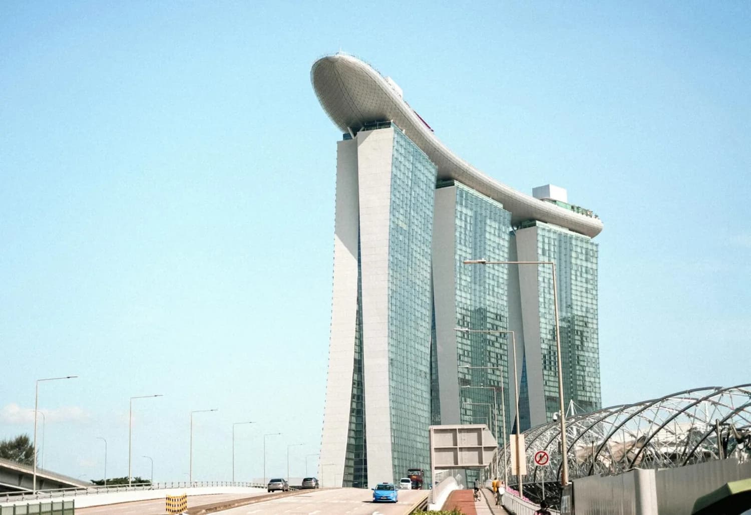 Marina Bay Sands Hotel in Singapur mit markanter Dachplattform, Straße im Vordergrund und Fußgängerbrücke
