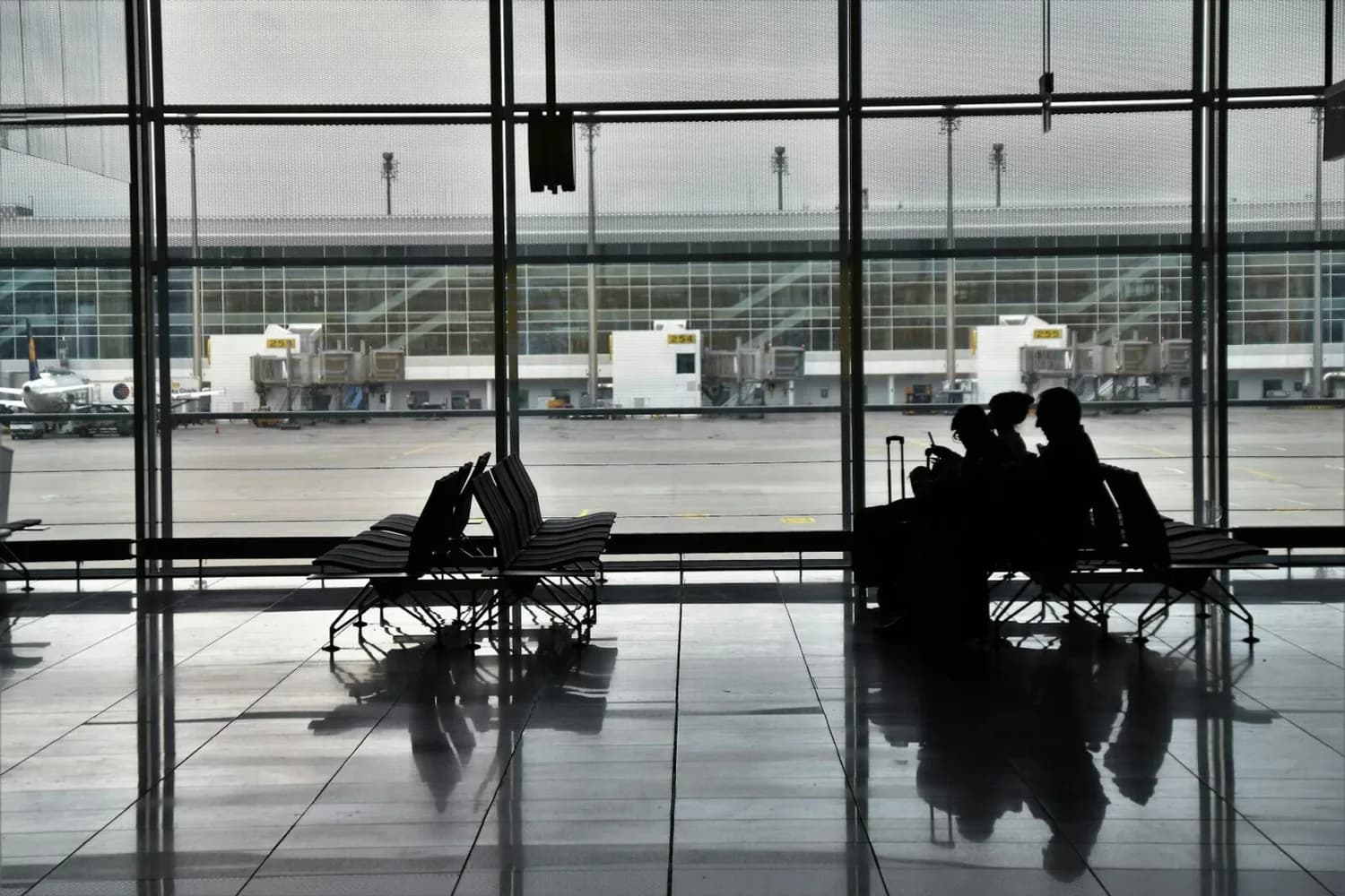 Wartebereich im Flughafen München mit Sitzreihen, silhouetten von wartenden Passagieren und Flugsteig sowie geparkten Flugzeugen hinter großen Glasfronten
