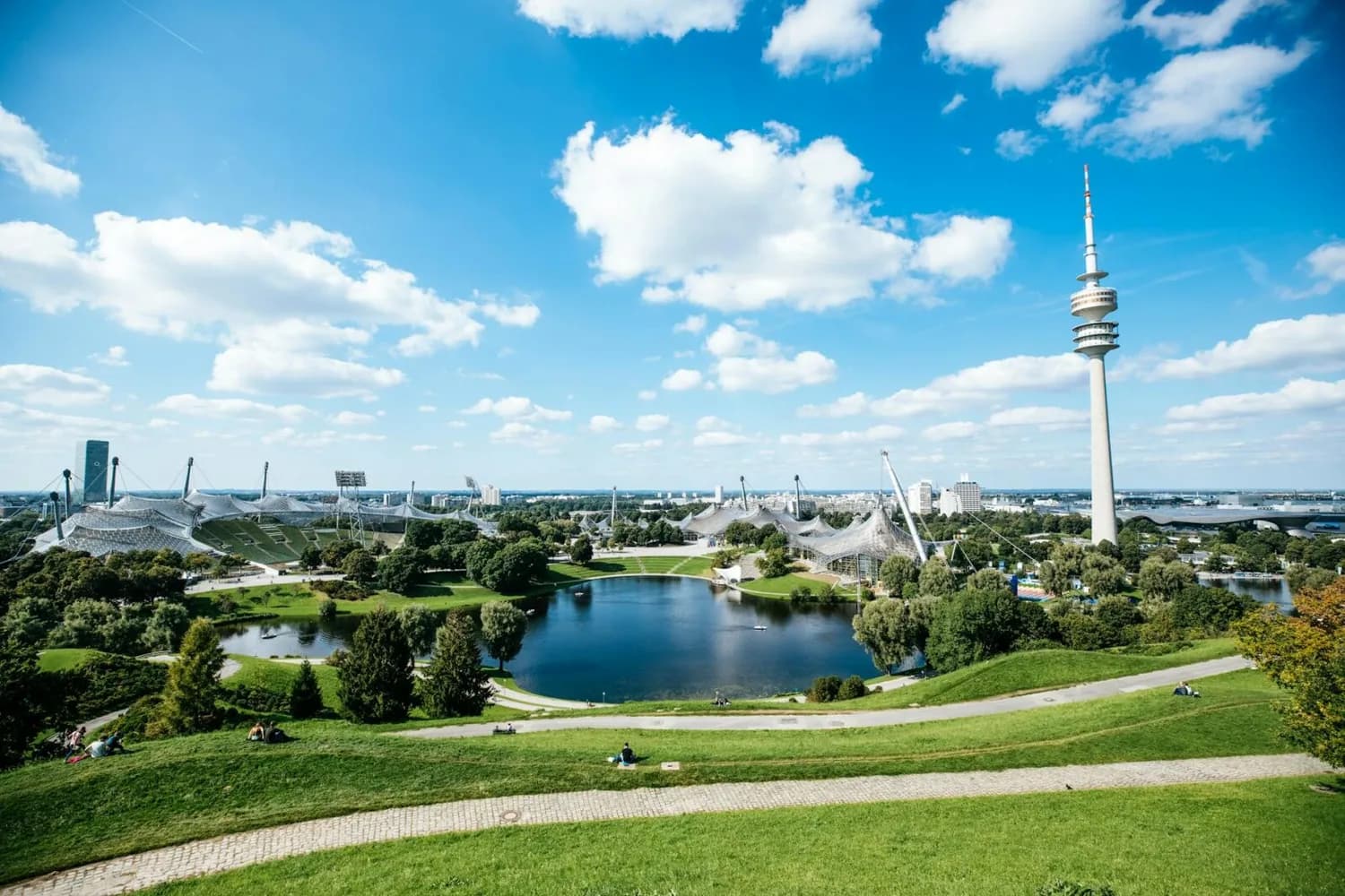 Panorama des Münchner Olympiaparks mit Olympiasee, Zeltdachstadion und Olympiaturm bei blauem Himmel