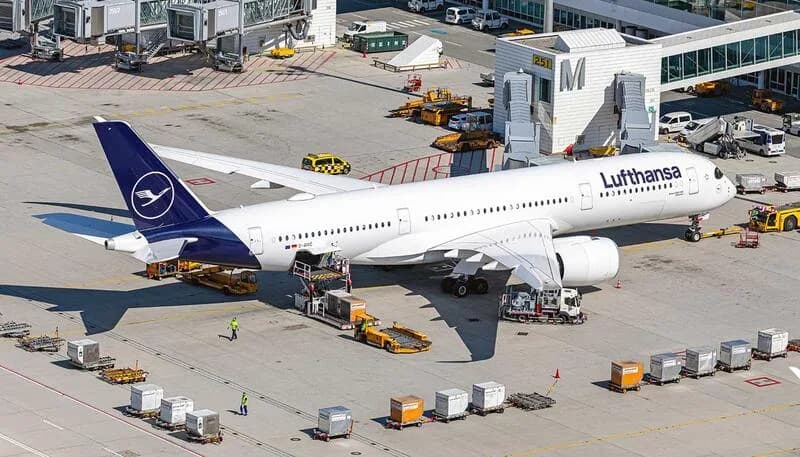 Lufthansa Passagierflugzeug A350 am Gate beim Be- und Entladen von Gepäck und Fracht, Bodenabfertigung mit Cateringfahrzeugen und Gepäckcontainern auf Flughafen-Gatebeltzonen - Bild (c) Flughafen München