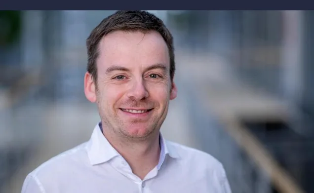 Mann lächelnd vor unscharfem Flughafeninterieur, Porträt von Sebastian Ibel am Flughafen München, Business-Kleidung, freundlicher Ausdruck