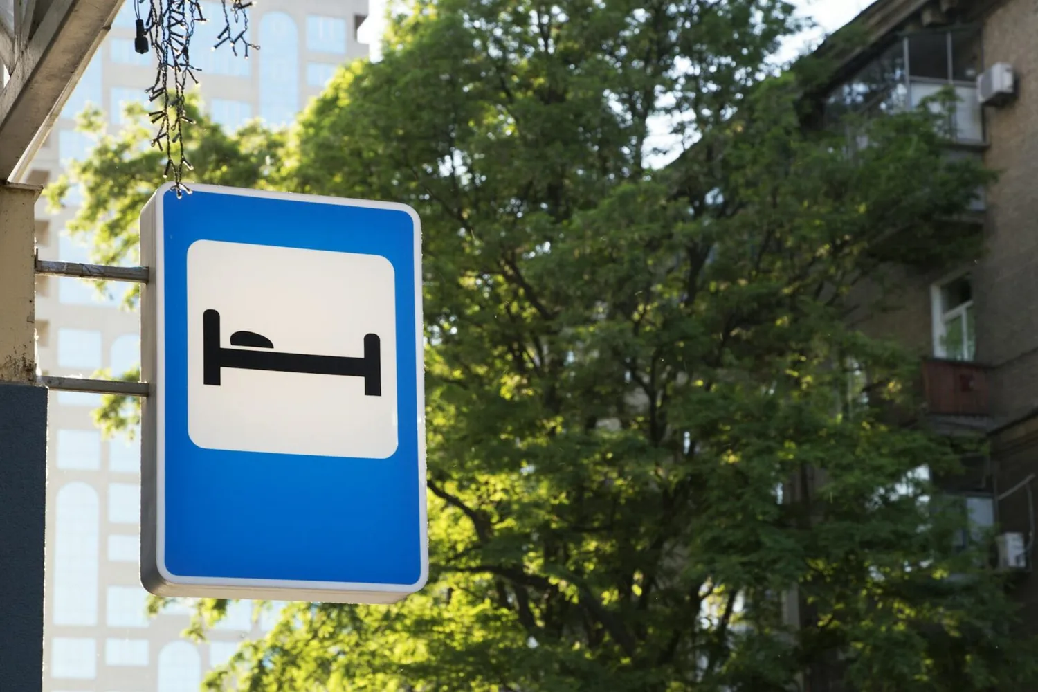 Symbolschild mit Bett für Hotelanzeige an Hausfassade, geeignet für Park-and-Sleep Park+Fly Flughafen München, blaues Verkehrszeichen vor grünen Bäumen