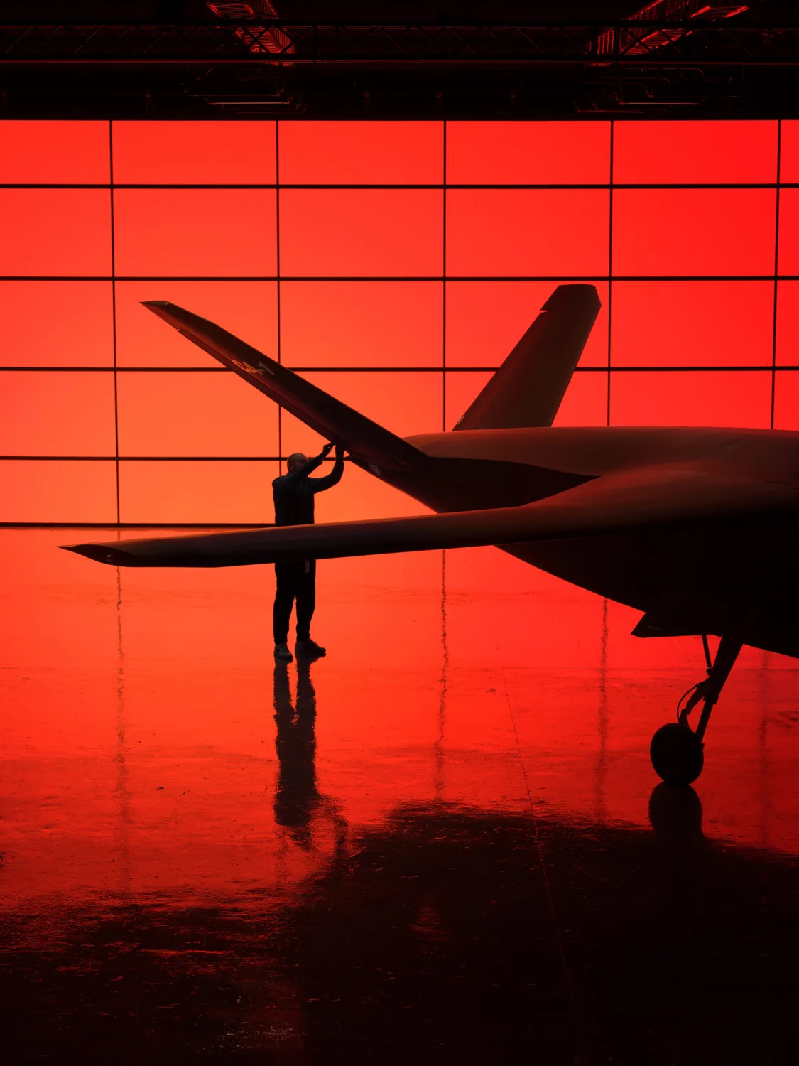 Silhouette einer Person, die an einem Flugzeugflügel in einem Hangar vor roter, quadratischer Hintergrundbeleuchtung arbeitet