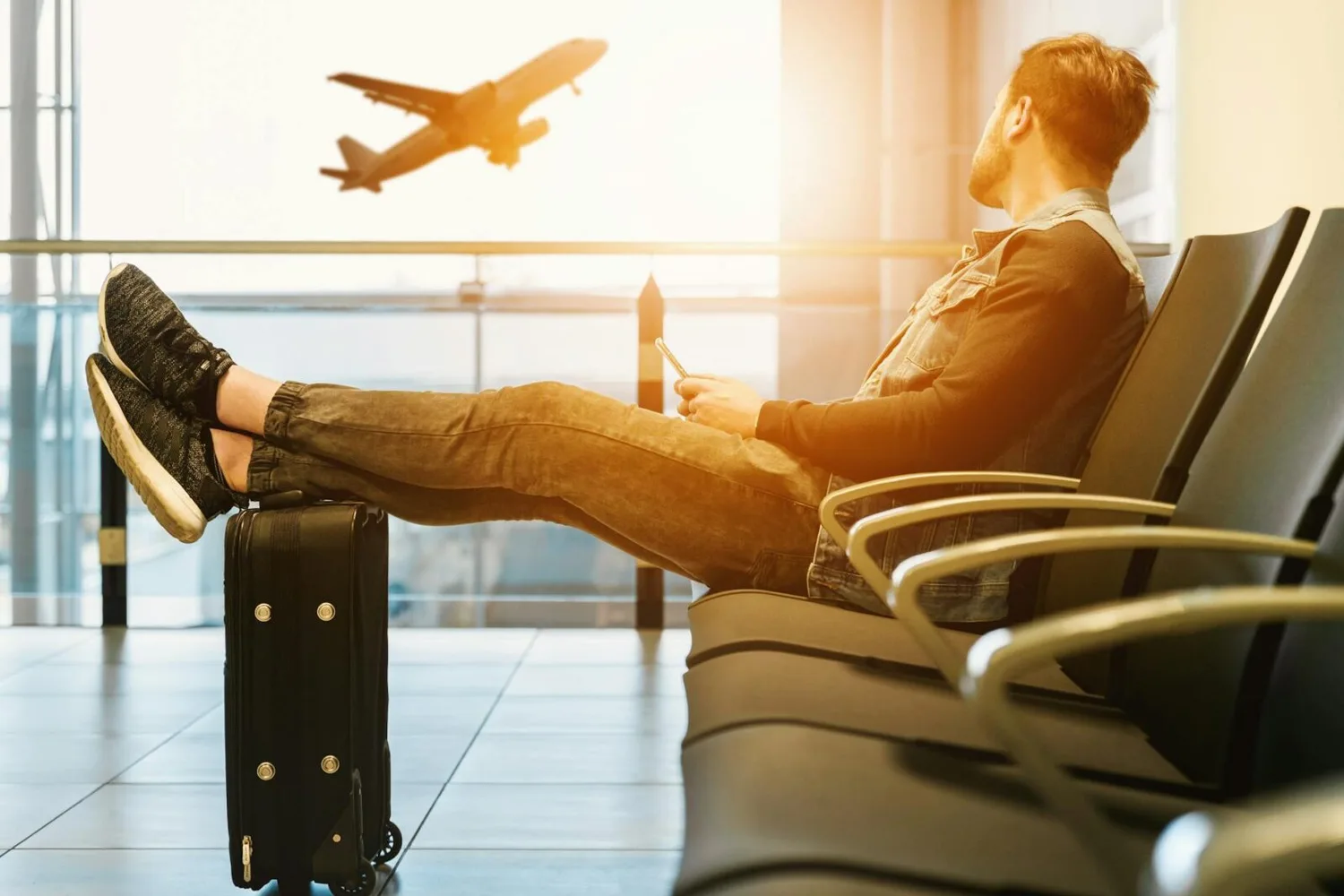 Mann entspannt im Flughafenwartebereich mit Koffer als Fußstütze, Flugzeug startet im Hintergrund, reisen ohne Gedränge, FJS-Reiseplanung