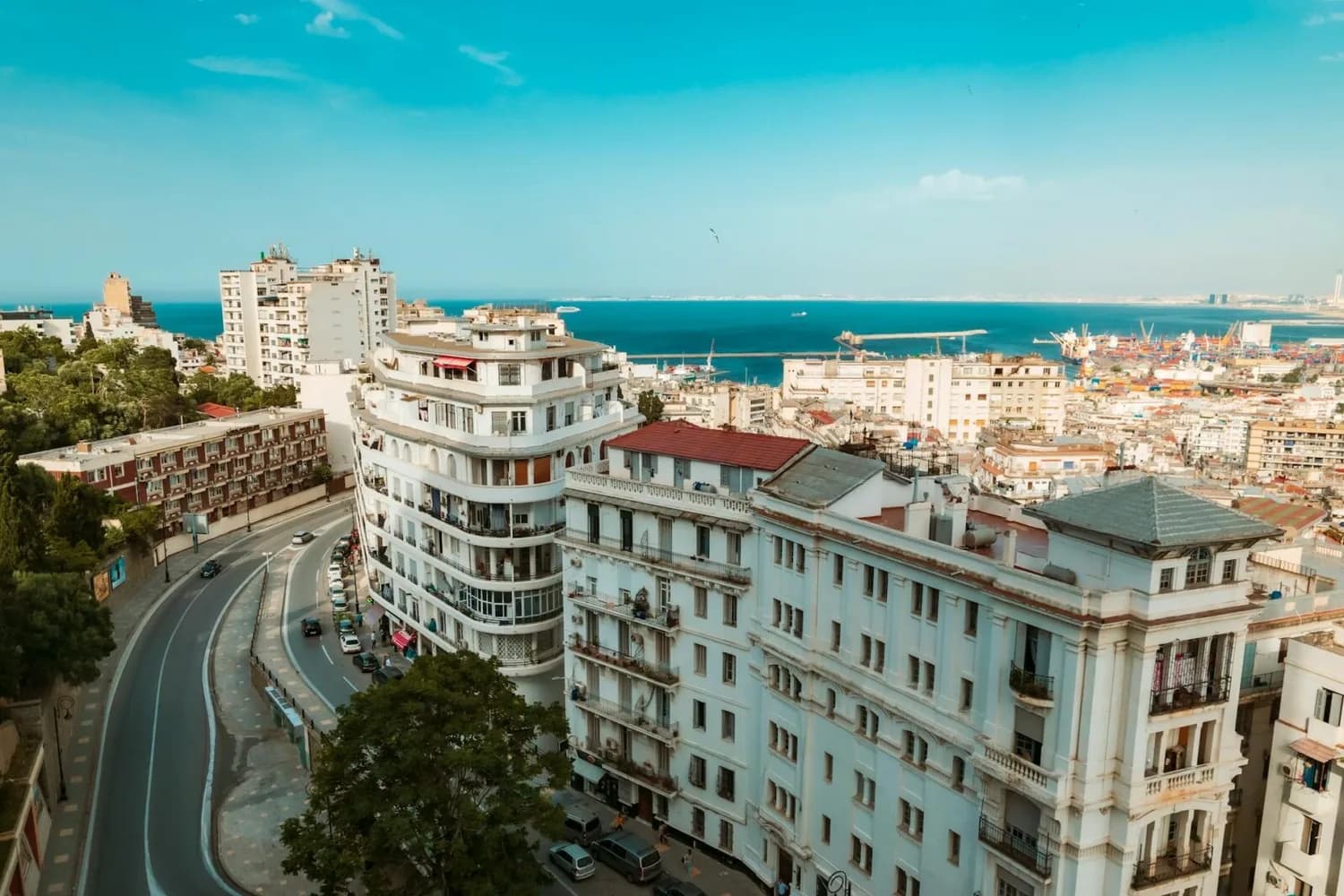 Blick über Algier mit weißer Küstenarchitektur, geschwungener Promenadenstraße und Hafen am Mittelmeer, Städtereise ParkSleepFly-Motiv