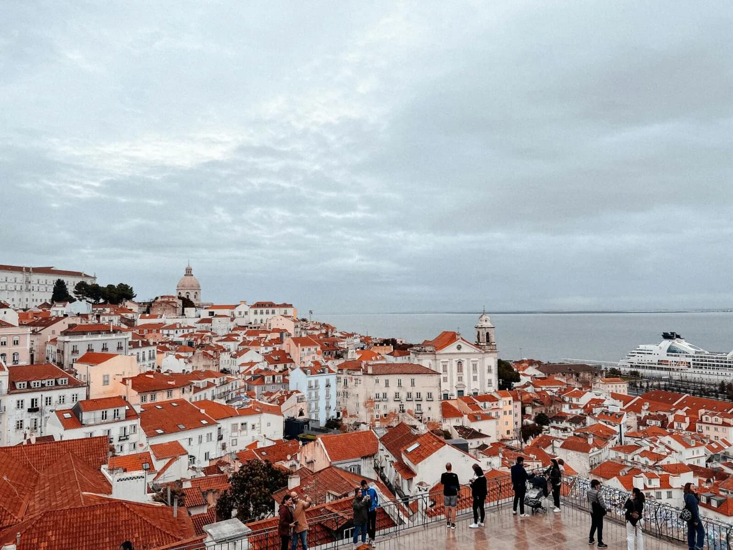 Panoramablick auf Lissabon mit roten Dachziegeln, historischen Gebäuden, Kirche und Tejo-Fluss im Hintergrund, Besucher an Aussichtsplattform