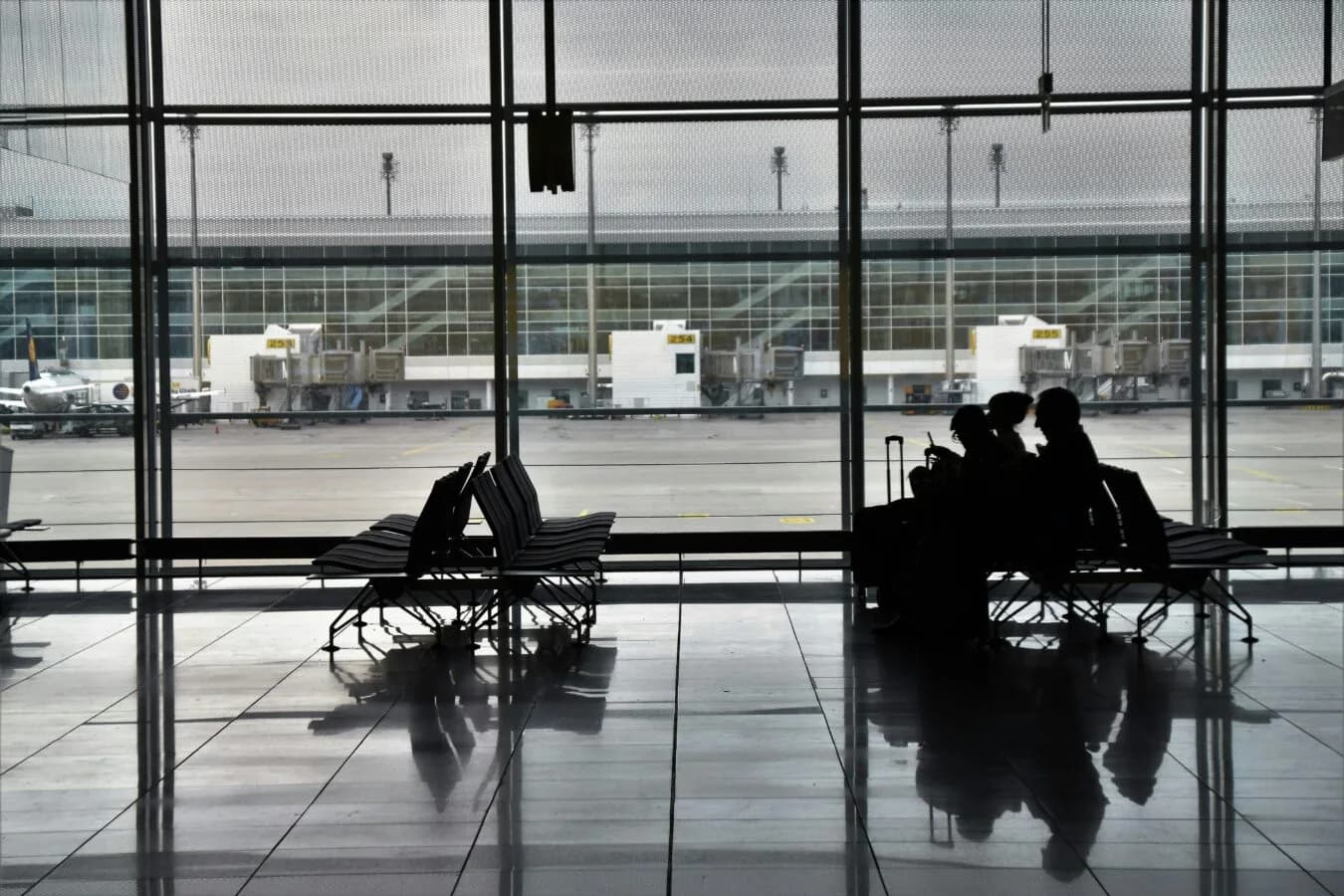 Wartebereich im Terminal mit Silhouetten von Reisenden vor großen Panoramafenstern und Flugsteigansichten am Flughafen München