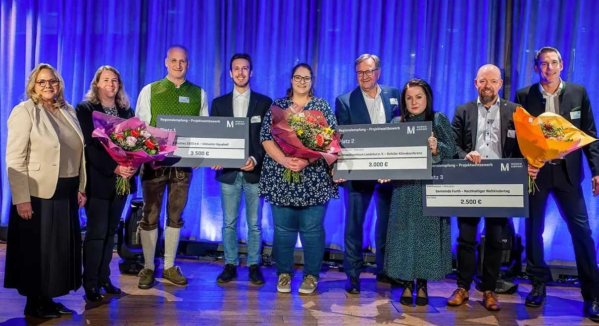 Preisträger mit Blumensträußen und großen Schecks auf Bühne bei Preisverleihung am Flughafen München vor blauem Vorhang