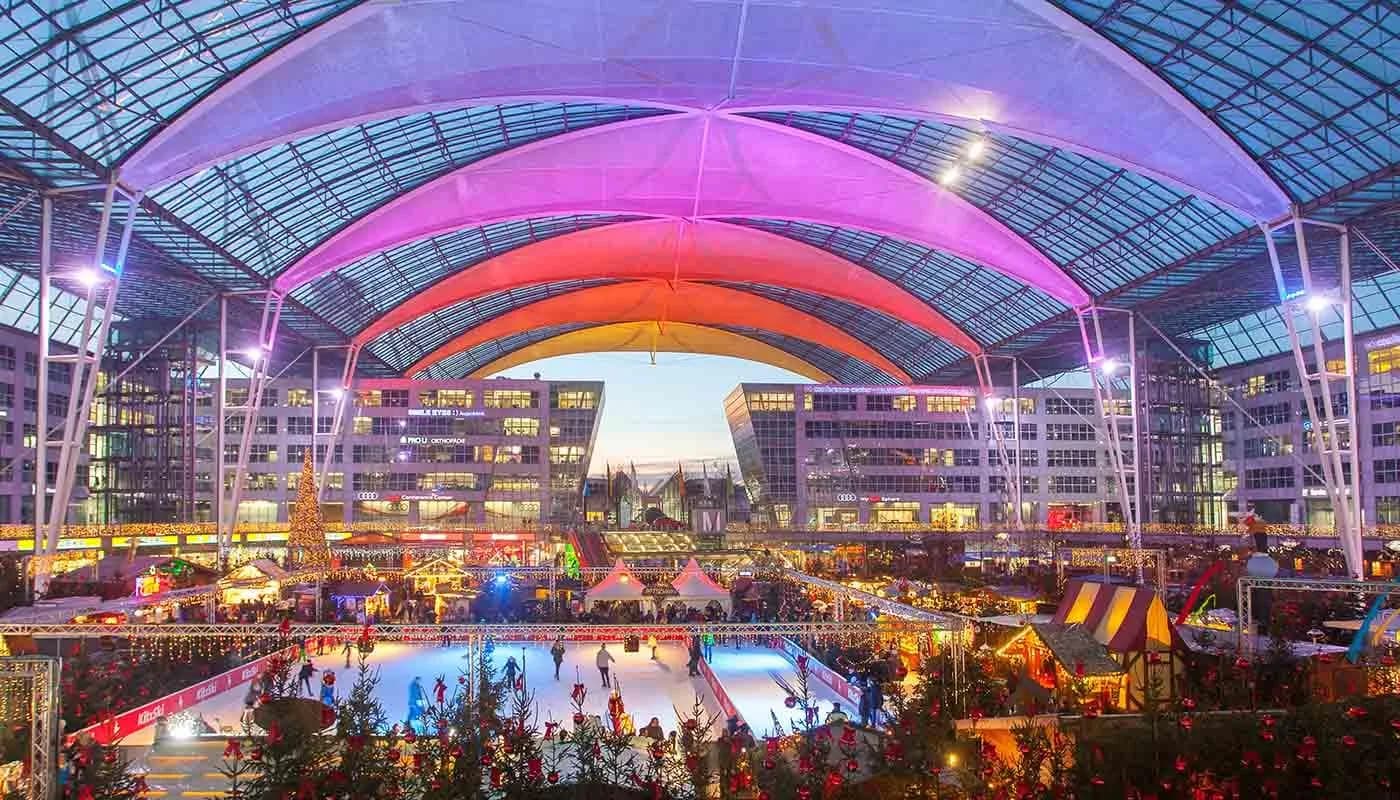 Überdachter Weihnachts- und Wintermarkt am Flughafen München mit Eislaufbahn, Lichtern, Marktständen und bunt beleuchteter Dachkonstruktion