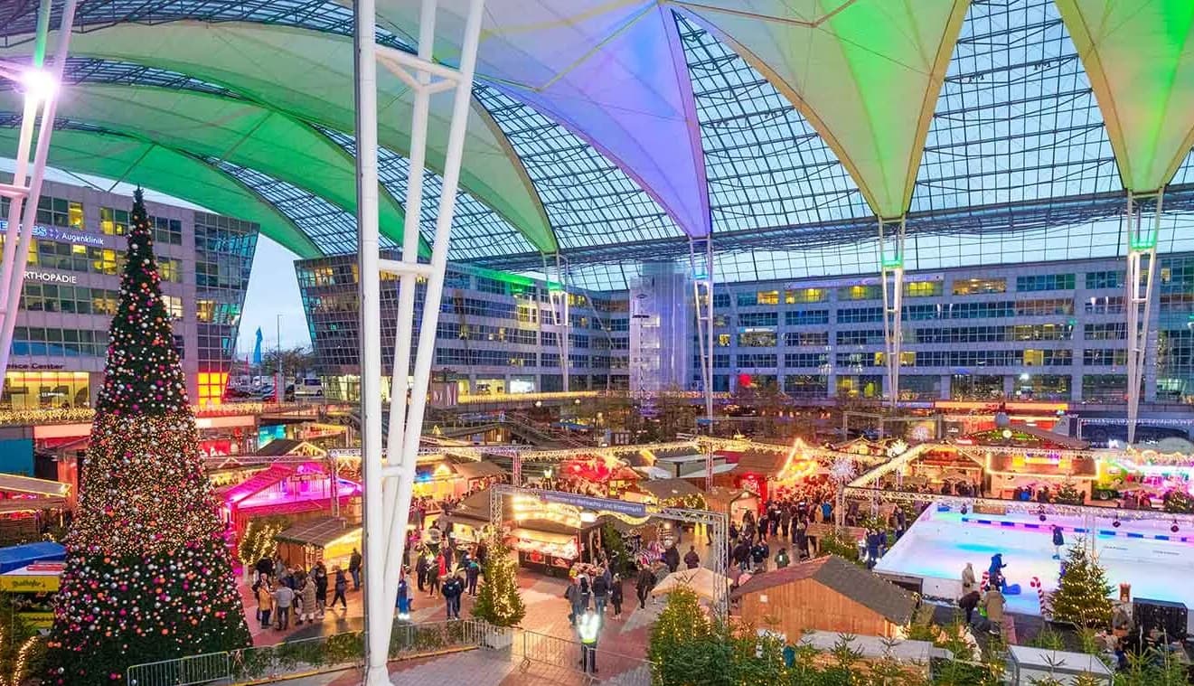 Überdachter Weihnachtsmarkt mit festlich beleuchteten Buden, großer geschmückter Tanne und Eisbahn im MAC Terminal des Flughafens München