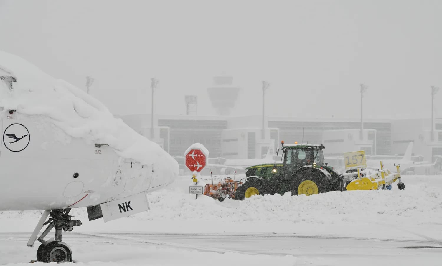 Schneebedeckte Flugzeugnase am Flughafen München und Traktor mit Räumschild im Winterdienst bei starkem Schneefall