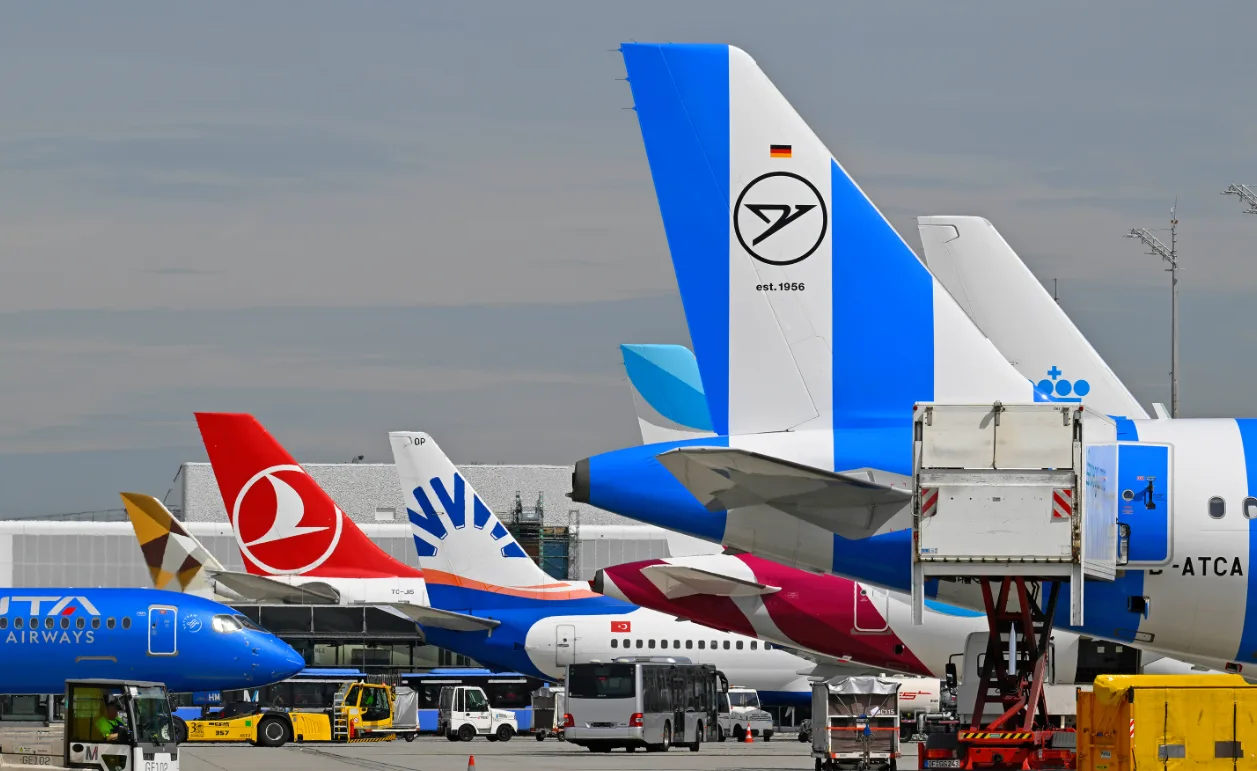 Reihenparkende Flugzeughecks mit bunten Leitwerken und Bodenservice am Flughafen München während Herbstferien