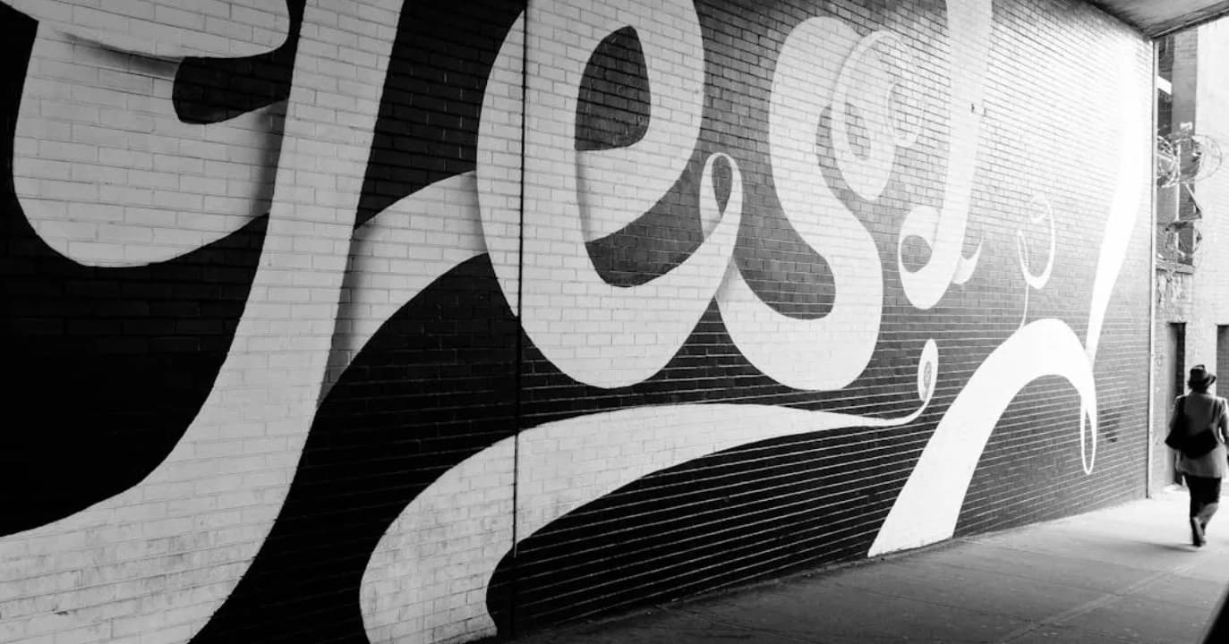Schwarz-weiß-Foto einer großen Wandgrafik mit geschwungenen weißen Formen auf dunklen Backsteinen und einer Person, die an der rechten Seite den Gehweg entlangläuft, urbanes Street-Art-Motiv parksleepfly muc