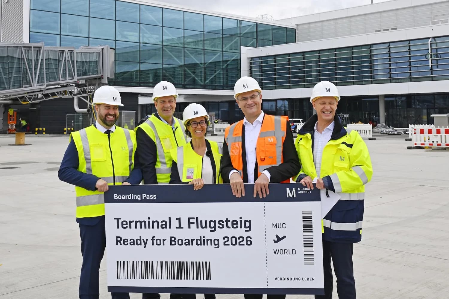Gruppenfoto von Bauleitern und Verantwortlichen in Schutzhelmen und Warnwesten vor dem Terminal 1 Erweiterungsbau am Flughafen München mit Passagierbrücke im Hintergrund