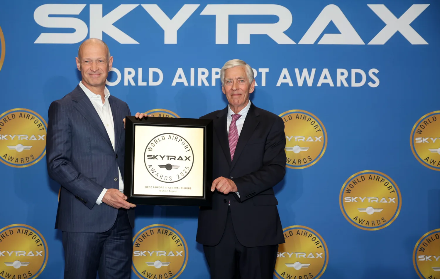 Zwei Männer in Anzügen halten bei den Skytrax World Airport Awards eine Auszeichnung für den Flughafen München vor blauem Messehintergrund mit Skytrax-Logo