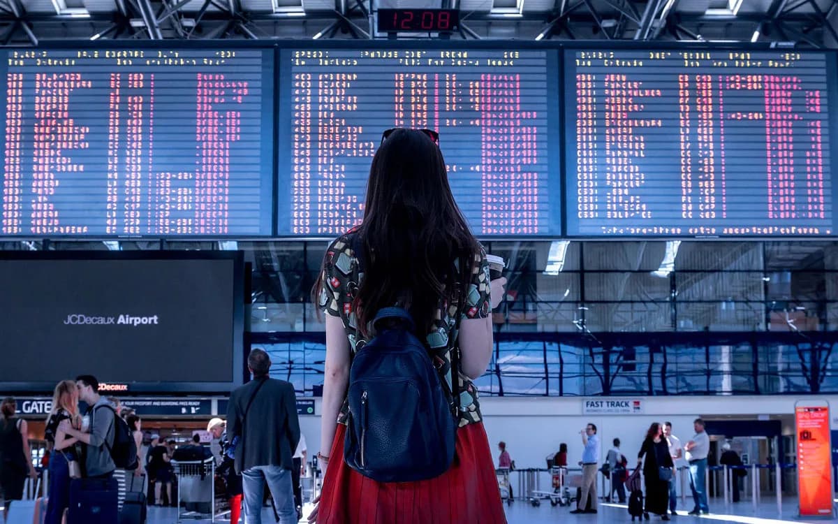 Reisende Frau mit Rucksack vor großer Anzeigetafel im Flughafen, Fluginformationen sichtbar, Szene mit Passagieren und Check-in-Bereich, addon fly cashback