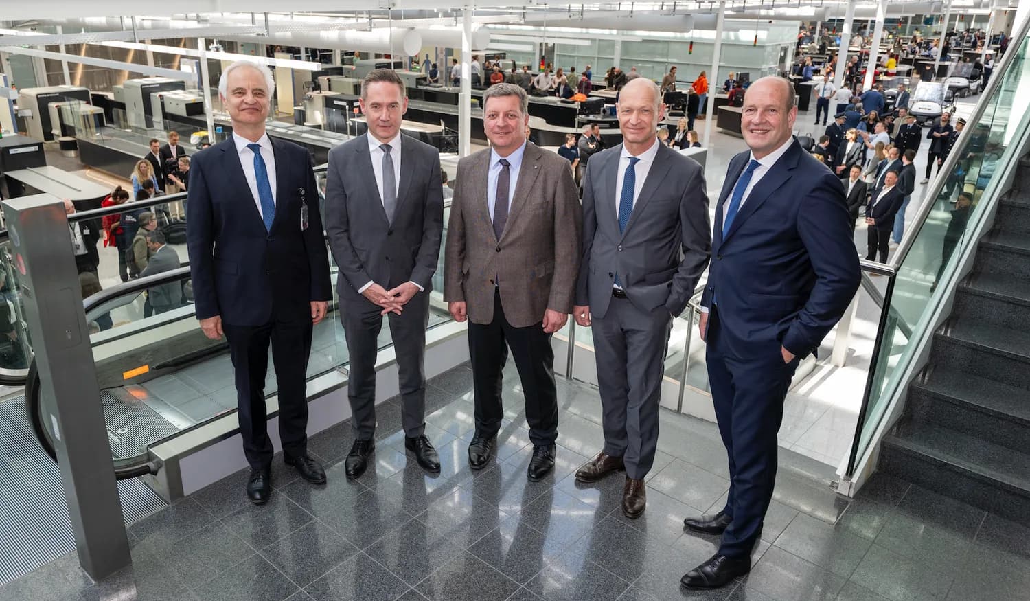 Fünf Geschäftsleute in Anzügen im Flughafenterminal vor moderner Sicherheitskontrolle mit Scannerbändern und Reisenden im Hintergrund am Flughafen München