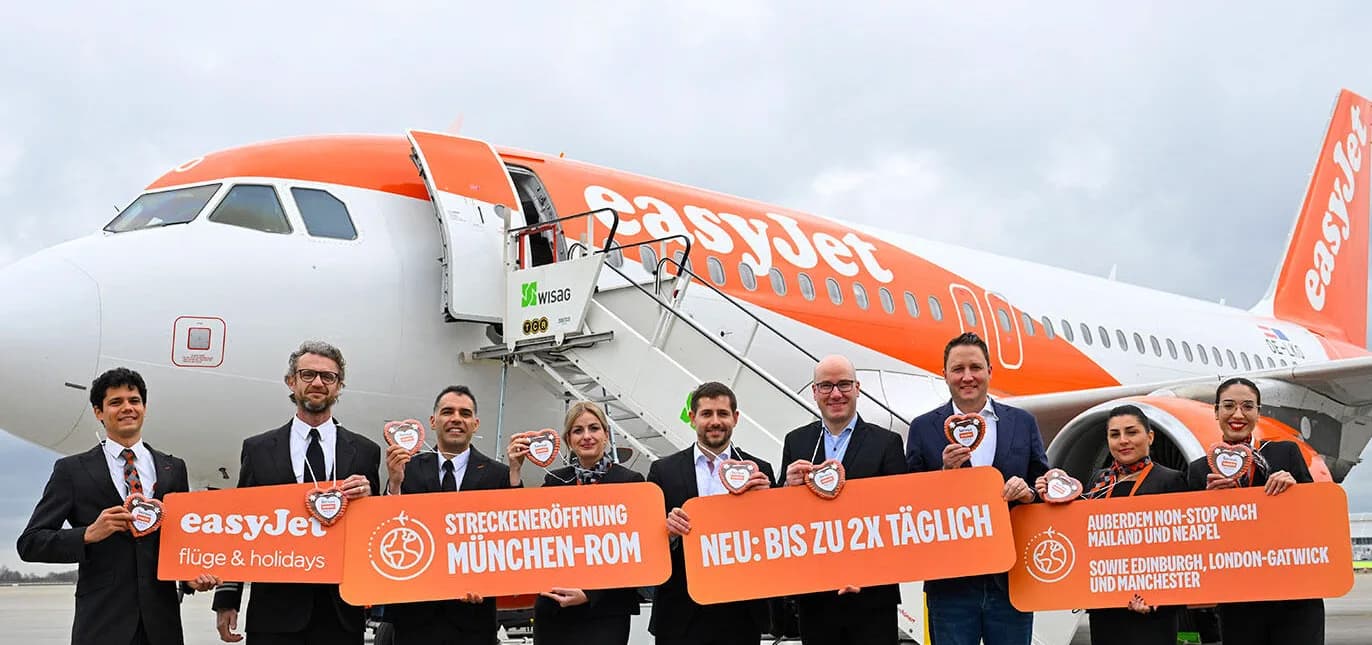 easyJet Passagierflugzeug am Flughafen München mit Crew und Veranstaltern, Banner zur Streckeneröffnung München–Rom und Ankündigung bis zu zweimal täglicher Flüge