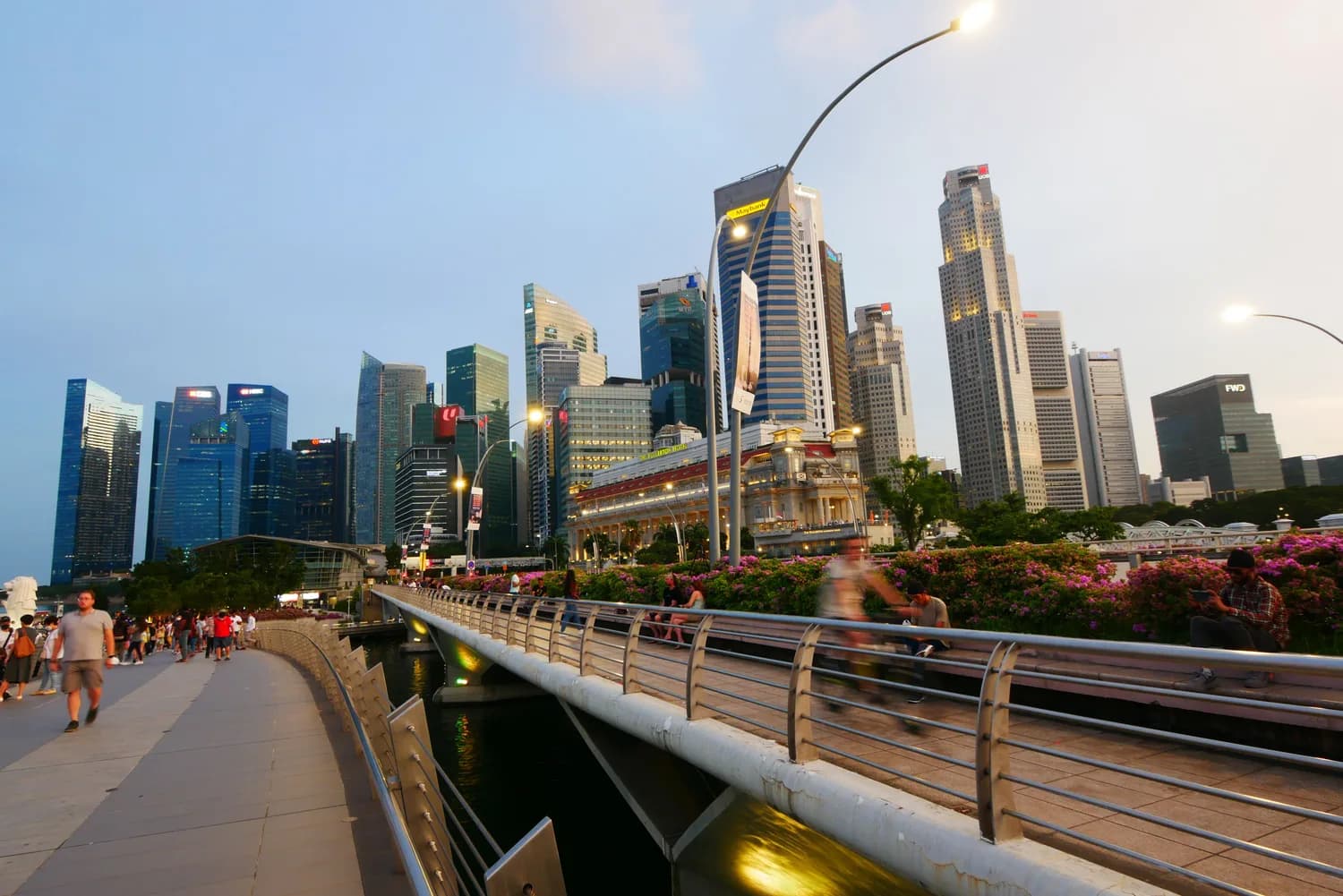 Abenddämmerung an der Marina Bay Promenade in Singapur mit Fußgängern, Radfahrern und moderner Skyline mit Wolkenkratzern im Hintergrund