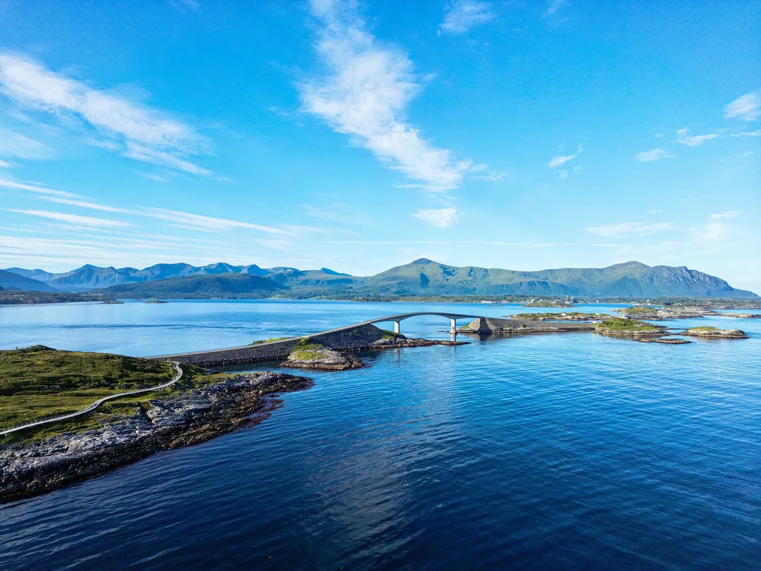 Atlantikstraße in Norwegen mit geschwungener Brücke, zerklüfteten Inseln und Bergen im Hintergrund bei klarem Himmel