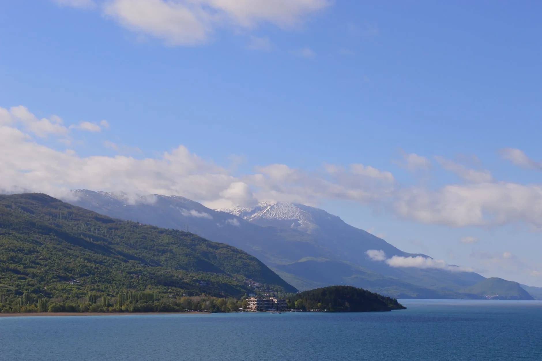 Weiter See vor bewaldeten Hügeln und schneebedeckten Bergen unter blauem Himmel mit Wolken