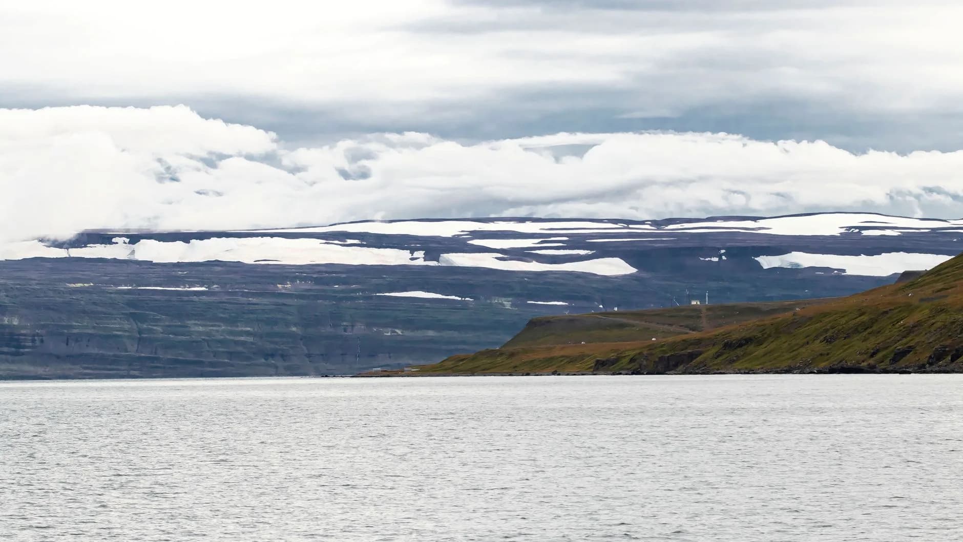 Fjordlandschaft mit schneebedeckten Felsformationen, grünen Hügeln und ruhigem Wasser unter bewölktem Himmel