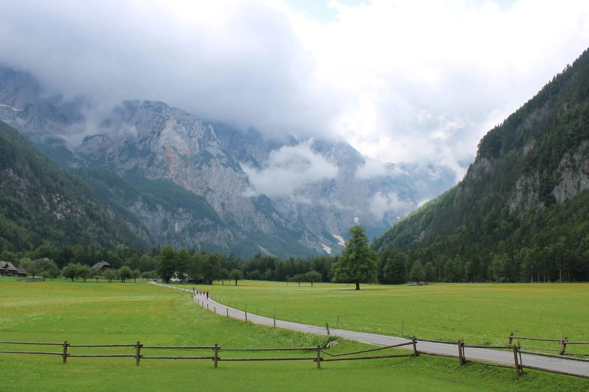 Weg durch grünes Bergtal mit Holzzaun, Wanderern und wolkenverhangenen Felsgipfeln im Hintergrund