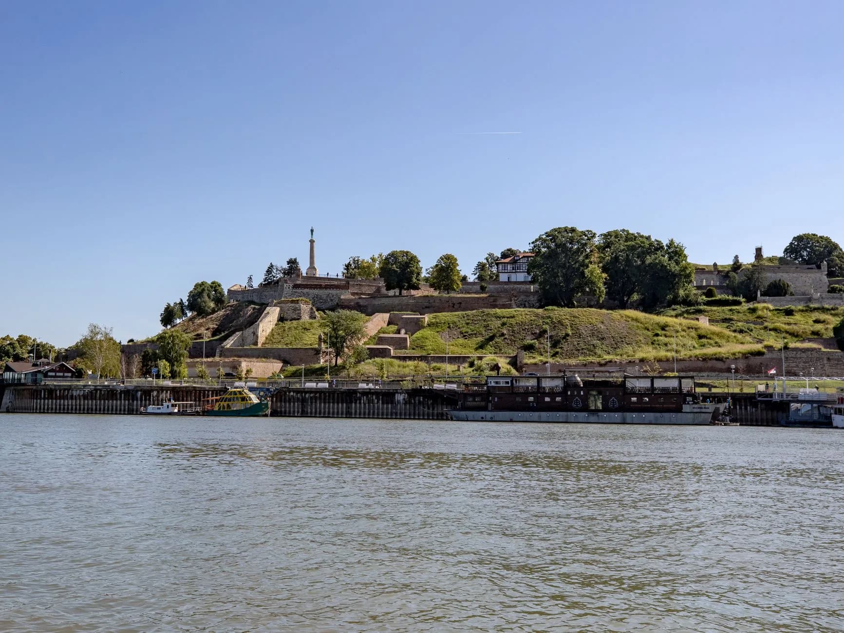 Festung auf einem Hügel über dem Fluss, mit Siegesdenkmal, Grünflächen und Hausbooten am Ufer