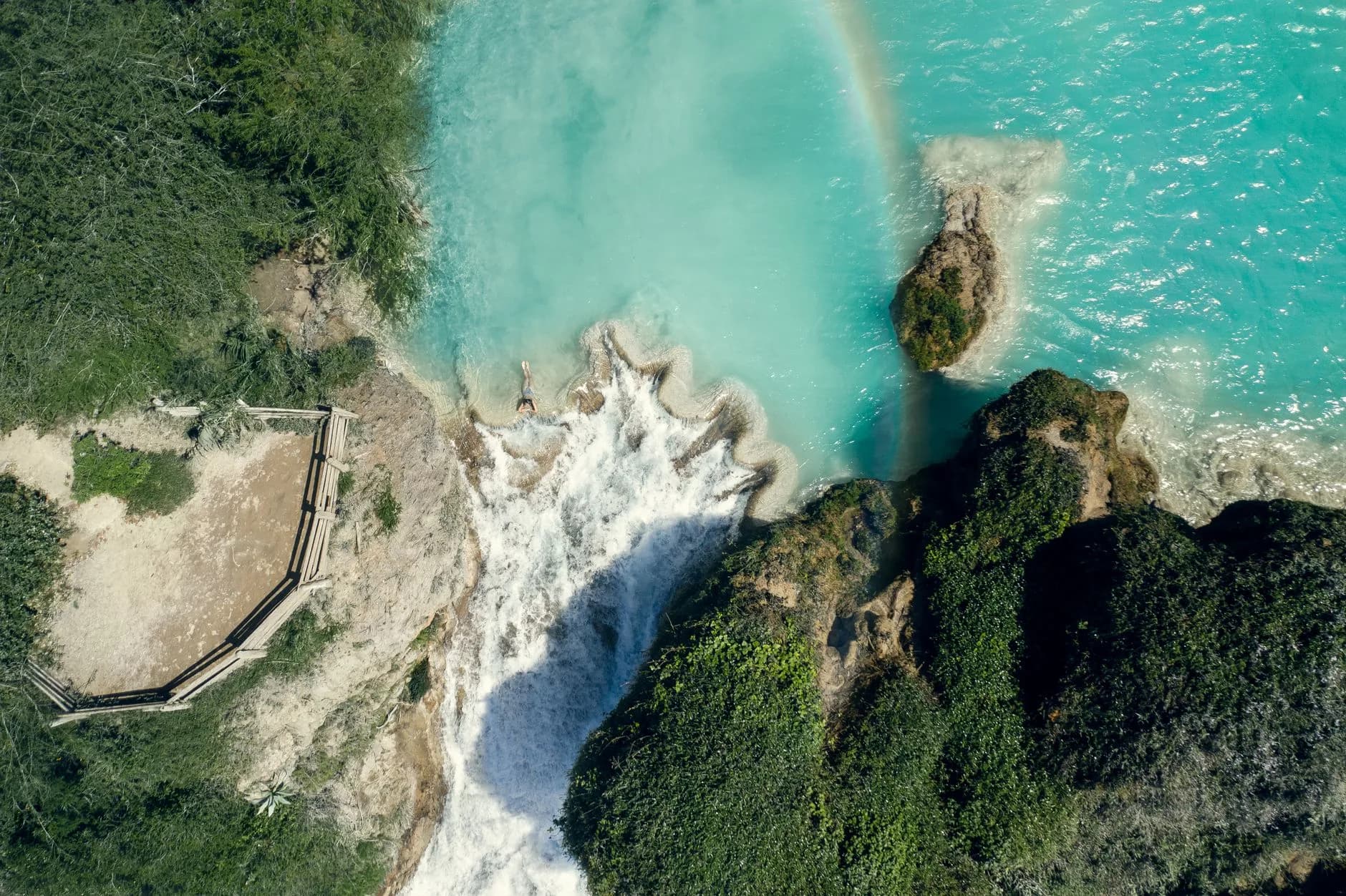 Luftaufnahme eines türkisfarbenen Wasserfalls an der Kante einer Lagune mit Regenbogen und einer schwimmenden Person
