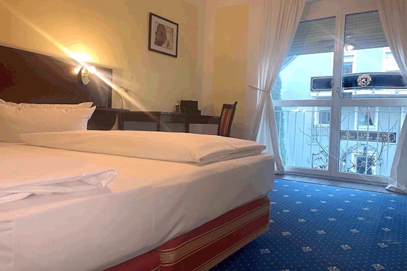 Zimmer mit großem Bett, Nachttischlampe, Schreibtisch, Stuhl und Balkonfenstern.