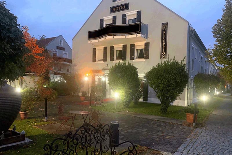 Eingang eines beleuchteten Hotels am Abend mit Vorgarten, Tischen, Gehweg und RECEPTION-Schild.