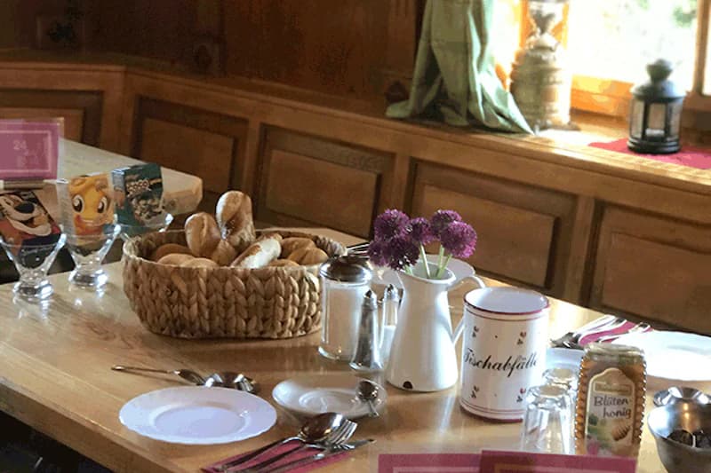 Gedeckter Holztisch mit Brötchenkorb, Tellern, Besteck, Vase mit lila Blumen und Glas Honig.