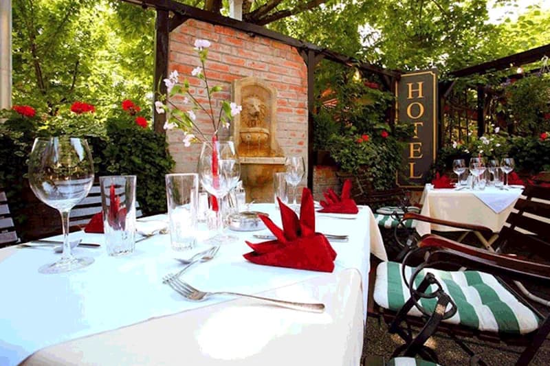 Hotelterrasse mit gedeckten Tischen, roten Servietten, Weingläsern, Backsteinbrunnen und HOTEL-Schild.