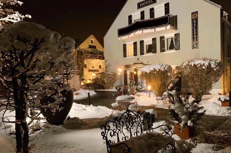 Verschneiter Hotelhof bei Nacht mit beleuchteter Fassade, Empfangsschild, Tischen und Metallbänken.