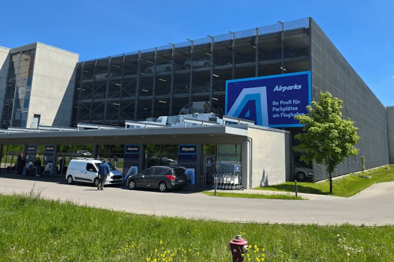 Mehrgeschossiges Parkhaus mit Eingangsüberdachung, Airparks-Schildern, parkenden Fahrzeugen und Personen im Vordergrund.