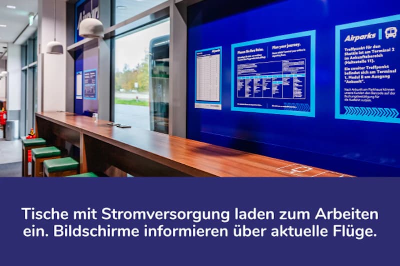 Innenraum mit langer Holztheke, Hockern, Steckdosen und blauen Infobildschirmen an der Fensterfront.