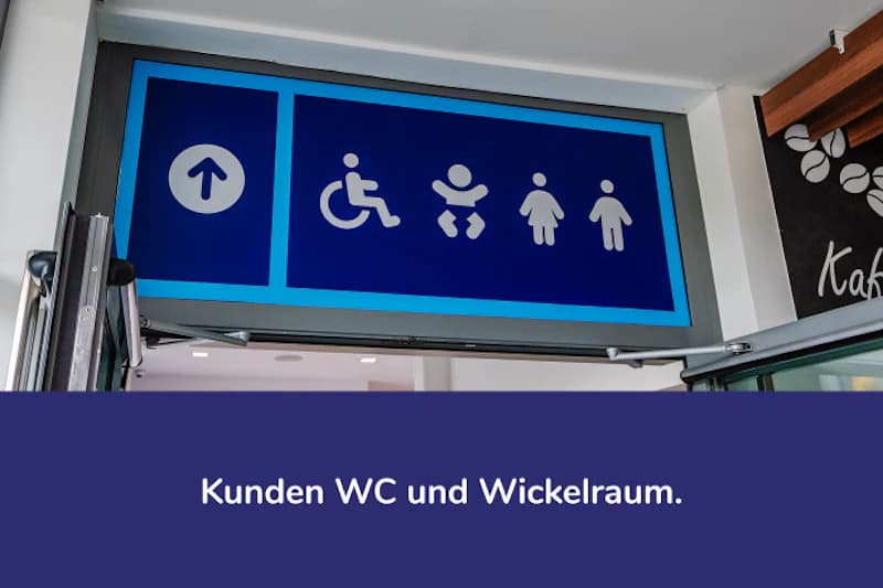 Blaues Schild über Durchgang mit Pfeil, Rollstuhl-, Wickel- und Damen- sowie Herrenpiktogramm.