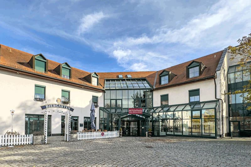 Dreigeschossiges Hotel mit Glas-Vorbau, überdachtem Eingang, Biergartenzugang und gepflastertem Vorplatz.
