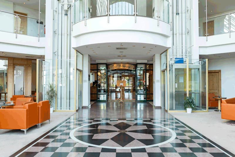 Große Hotelhalle mit gemustertem Marmorboden, Glasaufzügen links und rechts und orangefarbenen Sofas.