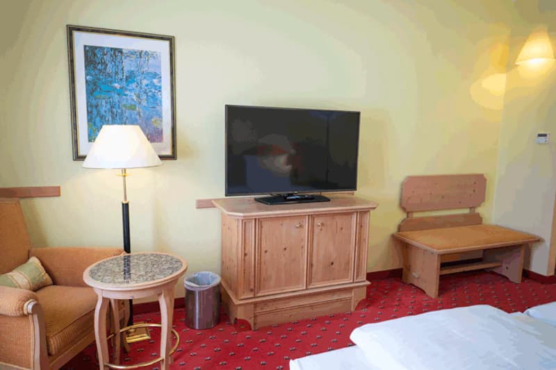 Hotelzimmer mit Fernseher auf Holzschrank, Sessel, Beistelltisch mit Lampe, Wandbild, Holzbänkchen und rotem Teppich.